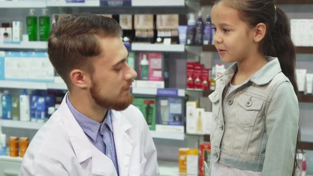 The little girl is talking to the chemist. He holds out his hand to her with pills. Then he tells her how to take these pills correctly. Mom and daughter are in the pharmacy