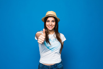 Obraz na płótnie Canvas Portrait of a smiling young girl wearing beach hat and showing thumbs up gesture isolated over blue