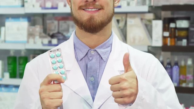 The pharmacist holds a packet of pills in his hand. With his other hand he shows a thumbs up. He smiles