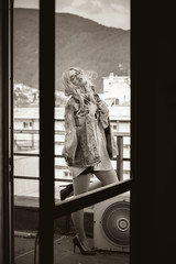 Blonde woman standing on a balcony wearing shorts, jean jacket  and high heels, cityscape as background