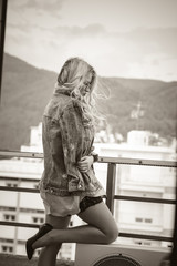 Blonde woman standing on a balcony wearing shorts, jean jacket  and high heels, cityscape as background