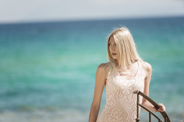 Fototapeta na wymiar Blonde woman in vintage knit dress with crochet patterns standing on stairs, ocean as background