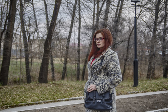 Stylish red hair lady in leopard coat and glasses with ladies handbag in autumn park
