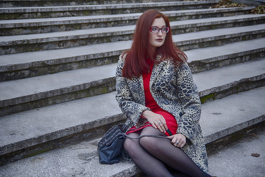 Stylish red hair lady in leopard coat, red dress and glasses with ladies handbag sitting on the concrete stairway