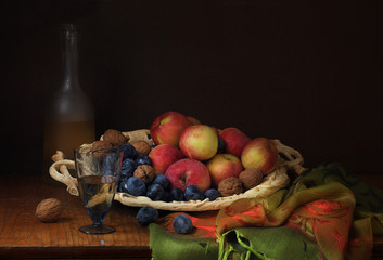 Apples, plums, walnuts on a carved dish, a bottle and a glass of wine, a silk tablecloth on an old wooden table. Classic still life style. Dark background.