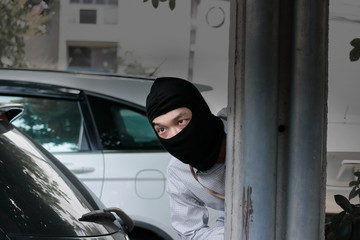 Masked burglar wearing a balaclava and sneaking behind concrete pole before the burglary in car park. Crime concept