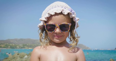Portrait of a beautiful little girl having fun on the sea, cute smiling in sunglasses, in a panama, background of sea blue water and rocks. Concept: children, childhood, summer, freedom, kids, baby.