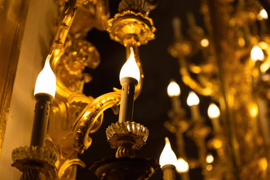 Close focus on bright warm candle with blurry scene of gold decoration on wall in dark environment.