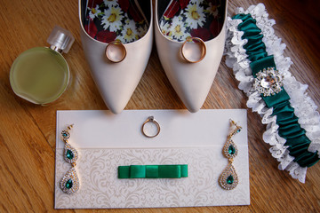 Wedding invitation near bridal jewelry with emerald, wedding rings, engagement ring, perfume bottle, garter and bridal shoes on high heels. Green wedding accessories for bride