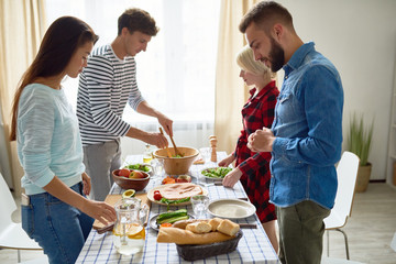Group of young people preparing dinner for festive celebration standing at big table together setting up food