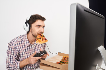 Portrait of young good-looking male gamer playing computer games with controller in headphones, holding pizza in mouth,looking in monitor with relaxed expression. © Cookie Studio