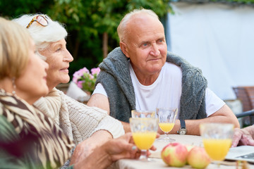Waist-up portrait of handsome senior man with deep blue eyes looking away while sitting at table with friends and celebrating momentous event at cozy small patio.