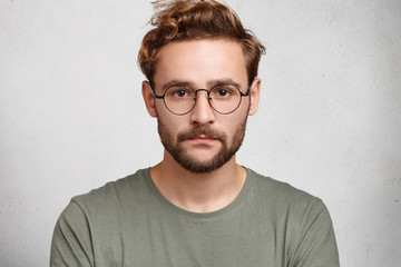 Headshot of serious bearded man with mustache and beard, wears round spectacles, has stylish hairdo, looks confidently, models in white studio, waits for partner. Young student going for classes