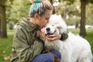 portrait of a young woman in a green coat and dreadlocks, kissing with his snow-white Samoyed dog...