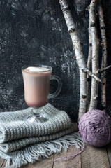 Homemade cocoa drink in glass cup on wool grey scarf. Cozy winter concept. Birch branches and wool knit ball on wooden table.  