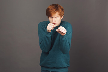 I'm not scared and going to fight you. Close up of funny red-haired male teenager in green sweater making fight pose, being scared but trying not to show this