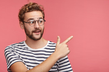 Excited bearded guy sees something unbelievable, indicates with fore finger at copy space for your advertisment or promotional text. Amazed young stylish male in spectacles points into distance
