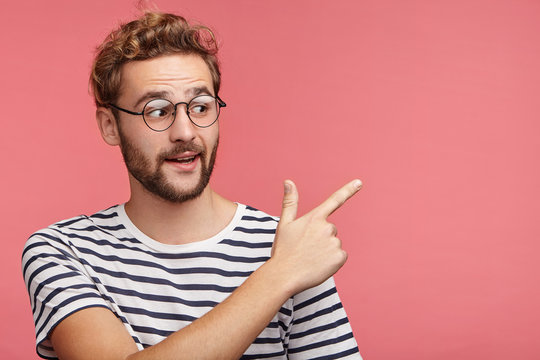 Excited bearded guy sees something unbelievable, indicates with fore finger at copy space for your advertisment or promotional text. Amazed young stylish male in spectacles points into distance