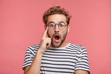 Studio shot of atractive fashionable hipster guy keeps mouth opened, being amazed by shocking news, isolated over pink background. Shocked stylish bearded man wonders, wears striped clothes.