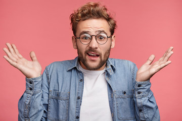 Waist up portrait of confused young man with trendy hairdo and stubble wears demin shirt and round spectacles, shrugs shoulders in bewilderment as makes difficult choice or decision in his life - obrazy, fototapety, plakaty