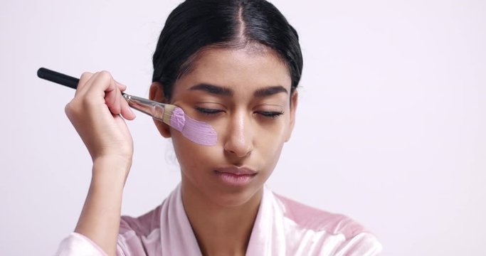 Pretty young girl with black hair and olive skin applying pink clay mask with a large makeup brush on white background
