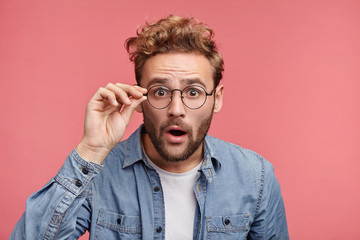 Terrified man dressed casually, stares through glasses, being surprised find out about his success. Male doesn`t believe his own luck, isolated over pink background. Emotions, people concept - obrazy, fototapety, plakaty