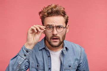 Puzzled emotional male with surprised expression looks through spectacles, tries to make out...