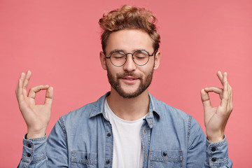 Relaxed carefree male student meditates against pink background, tries to relax after long hours of studying, concentrates on something, isolated. People, rest, relax, meditation, lifestyle concept