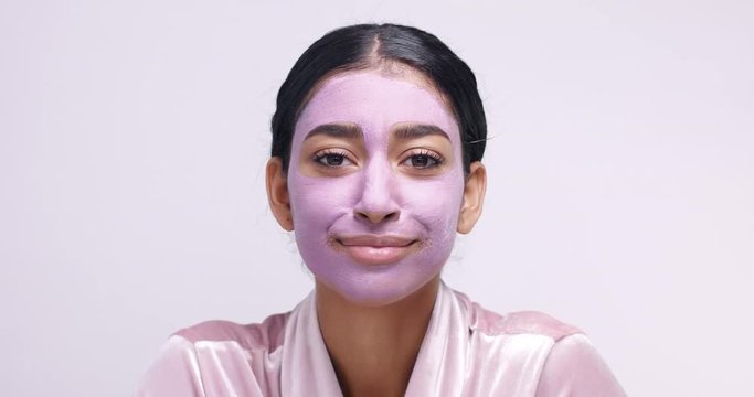 Pretty Middle Eastern young female model in pink clay cleaning mask making funny faces isolated on white