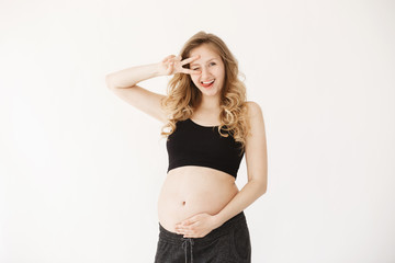 Isolated portrait of joyful young european pregnant woman with blond wavy hair in comfy clothes showing tongue, winking, making peace sign with hand, holding belly, having fun during photo session.