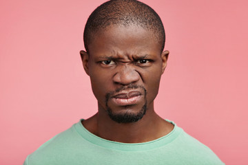 Displeased discontent African American male frowns face, dislikes something, looks with disgusting expression, isolated ovr pink background. Attractive unhappy young man isolated over pink background