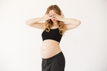 Naklejka premium Portrait of funny attractive european mother with blond curly hair in black sport suit with opened pregnant belly, clothing eyes with hands, having surprised expression. Pregnant woman playing with