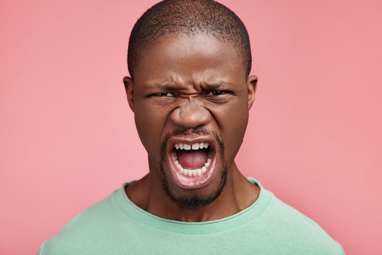 Furious African American male screams in despair, opens mouth widely, being outraged because of bad news, frustrated and discontent. Aggressive dark skinned man expresses madness, yells in anger