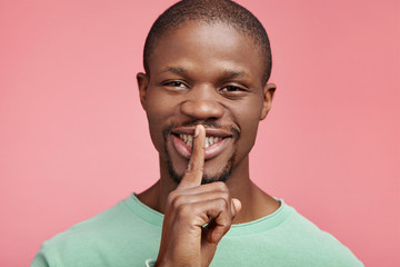 Happy dark skinned male keeps fore finger on lips, makes silence gesture, asks not to make noise or...