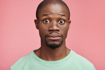 Bug eyed man with dark healthy skin, has full lips, stares through glasses, being terrified to know that he was dissmised for absence from work. Amazed scared Afro American man stand against pink wall