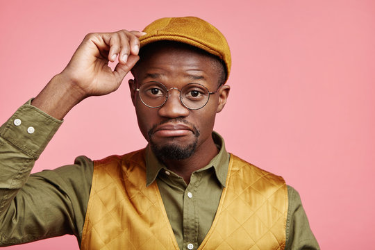 Portrait of hesitant stylish young hipster male with dark skin wears fashionable cap, shirt and vest, curves lips, has puzzled look, being uncertain, makes decision, isolated over pink background