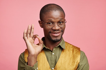 Horizontal portrait of dark skinned middle aged male with happy look, shows ok sign, gestures indoors, says: I have done it, everything is just fine. African male approves someone`s actions.