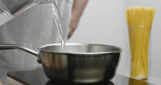 Chef in gray apron pours water to a large stainless steel pot and mixes with a wooden spatula on white background