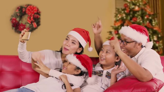 Cheerful family use a smartphone and making video call on Christmas day while wearing Santa hat on the sofa with Christmas tree background