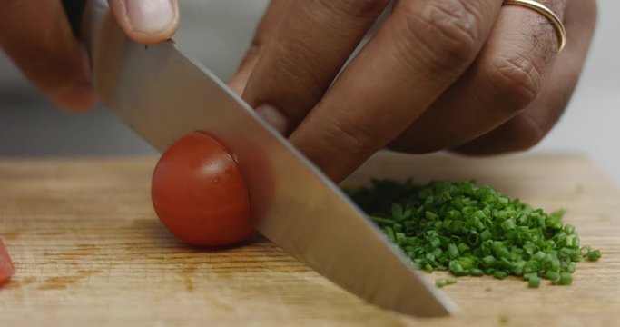 Dark skinned male cook cuts cherry tomatoes in half on a wooden chopping board next to chopped chives