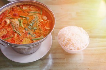 Popular Thai traditional spicy soup with shrimp or seafood call as “Tom Yum Kung” in Thai language in hot pot with a bowl of jasmine white rice on wooden table