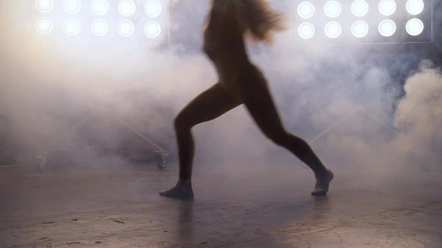 a young woman of pleasant appearance dances on stage with lighting projectors in the background. the girl professional dance performs the put number in a sty hip hop. A smoke machine works and a man