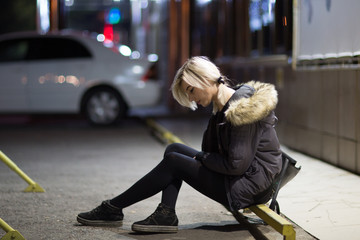 Tired depressed drunk young blonde woman sitting near wall in night city lights bokeh