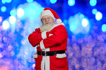 Brightful photo of realistic Santa Claus. Ola Santa Claus with crossed arms standing on blue shiny background. Santa Claus and New Year celebration.