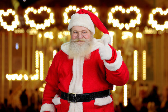 Santa Claus giving thumb up. Cheerful Santa Claus giving thumb up gesture on New Year festive lights and garlands.