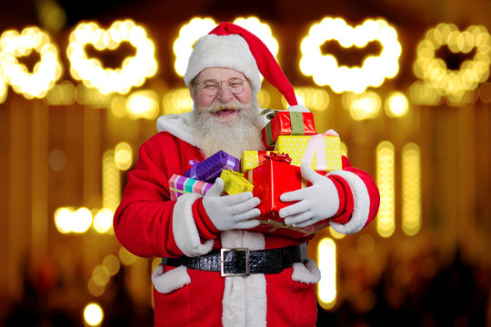 Santa Claus holding boxes with gifts. Smiling Santa Claus holding a pile of gift boxes against light design shimmering on dark.
