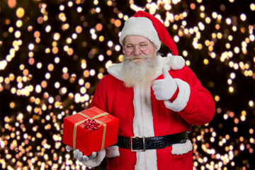 Santa with present giving thumb up. Happy Santa Claus holding red gift box and giving thumb up on New Year lights background.
