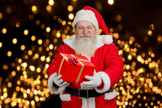 Santa Claus offering beautiful gift box. Smiling old Santa Claus holding present box on festive lights background. New Year and Christmas holiday concept.