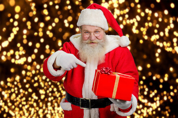 Santa Claus holding beautiful red box. Authentic Santa Claus holding red gift box and pointing on it with his index finger, festive lights background. Happy santa Claus with present box.