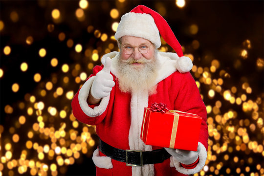 Santa with gift giving thumb up. Happy santa Claus in glasses holding red box with present and gesturing thumb up on New Yewr lights background.